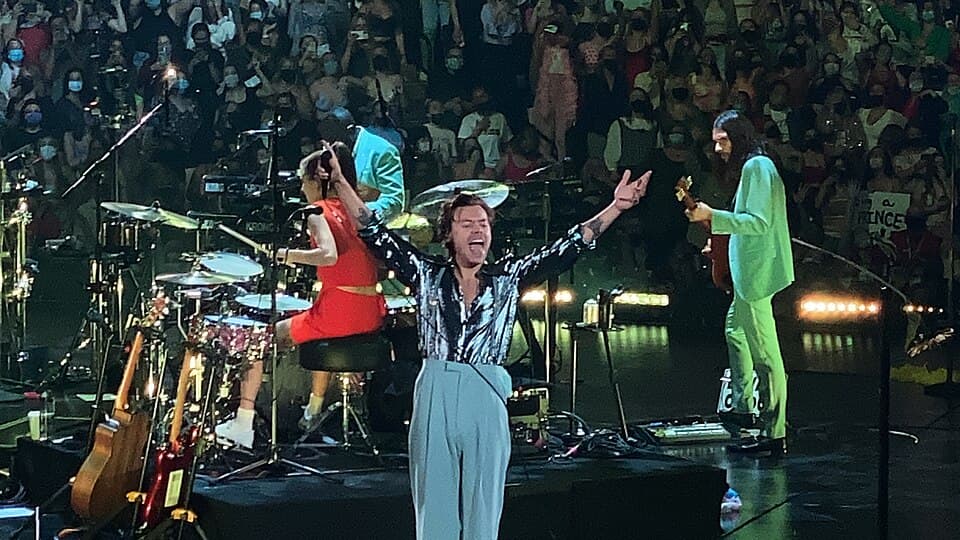 Harry Styles on stage at Xcel Energy Center, bathed in colorful stage lighting with an enthusiastic crowd surrounding the main floor