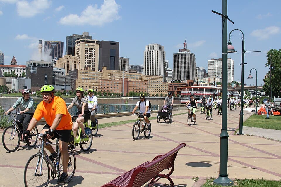 Cyclists riding along the Mississippi riverfront in Saint Paul at a Nice Ride kickoff event, with the downtown skyline visible across the water